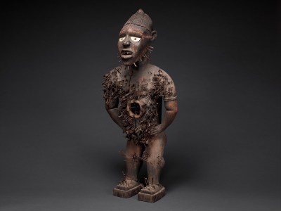 A wooden figure with its hands at its sides and a hole in its stomach. Metal nail-like elements are driven through the center.