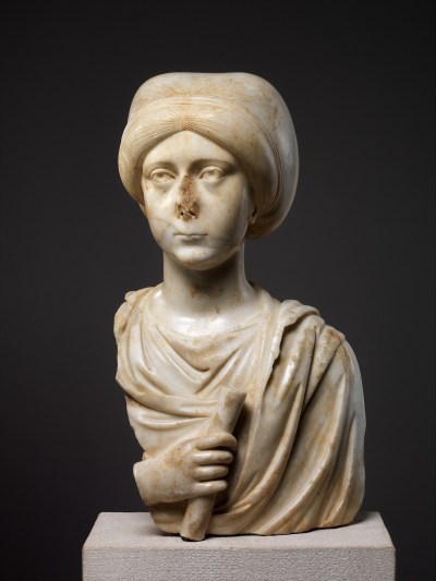 A bust of a woman wrapped in a shawl-like garment and a turban. Her nose and one of her shoulders are broken off, and she holds a scroll in one hand.