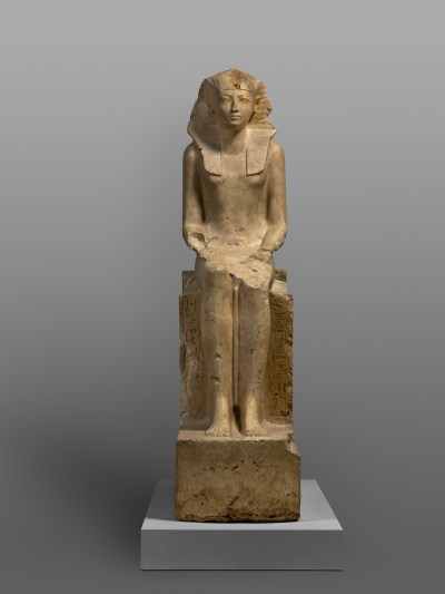 A statue of a seated woman wearing a headdress that partly covers her bared breasts.