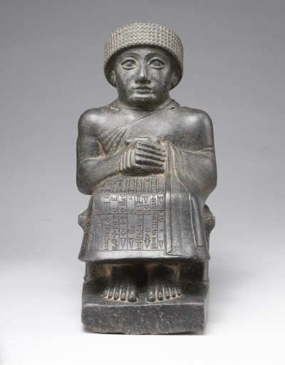 A statue of a man sitting with his hands clasped. He wears a toga-like garment that has Sumerian characters carved into it.