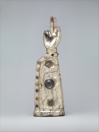 A metal sculpture of a raised arm with three fingers pointing up. Jewels are inset in the sleeve containing the arm.