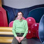A man in a green shirt sits in front of a jewelry painting.