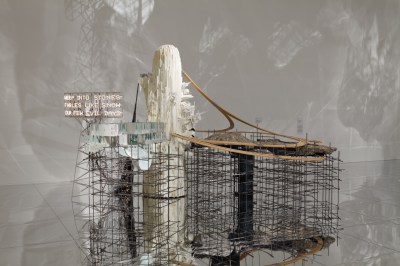 An installation composed of a metal grate and wood elements, along with a sign reading 'WEEP INTO STONES FABLES LIKE SNOW OUR FEW EVIL DAYS.'