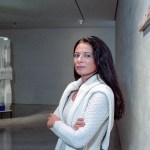 A woman in a white knitted sweater stands with her arms crossed next to an abstract sculpture.