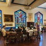 Group of people sitting on tables in a room with religious stained glass windows.