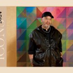 A man in a black vest wearing a black shirt in a black hat standing in front of a multi-colored abstract painting in the work.