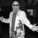 Black and white photo of an old bearded man smiling with arms, wearing a white suit with face printed on it