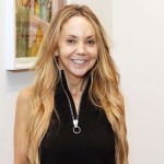 Portrait of a smiling white woman in black top with long zipper extending from the middle. Behind her is an abstract painting.