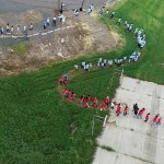 Aerial drone shooting of elementary school students of multiple ages, curved lines along the river shape. Students wear matching T-shirts in turquoise, sky blue or red with project logos.