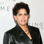 A woman wearing a black leather jacket posing in front of a white wall. and necklace.