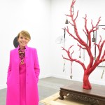 A woman in a purple coat stands next to a mangrove sculpture with various objects hanging on it.