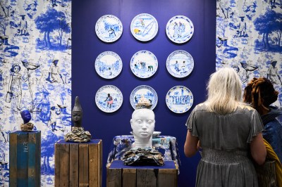 An installation featuring ceramic busts on plinths with shipping paletts covering them. Behind is a 3x3 grid of ceramic plates and wall paper.