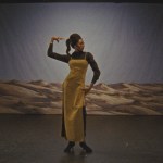 A woman in mustard colored clothes holds one of her hands. Behind her is a background with images of arid landscapes.