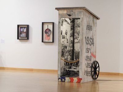 Two mixed media artworks hang on the wall behind the sculpture, resembling an outhouse that combines with photographs from the Civil Rights Movement. 