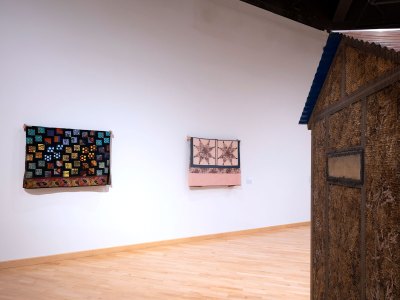 Two quilts hung on the wall. On the left is a partial view of the sculpture resembling the shed. 