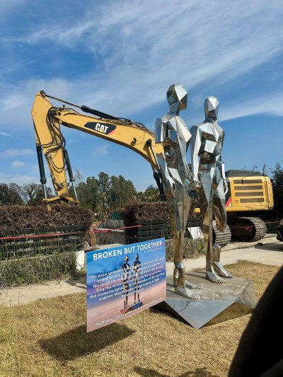 A reflective metal sculpture made of various polygons that resemble two standing figures holding hands. A crane sifting through a burned home is seen in the background. 