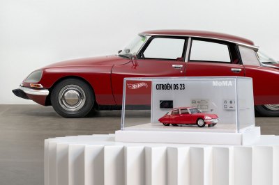 A replica of a red sports car sits on a pedestal in front of the car that inspired it.