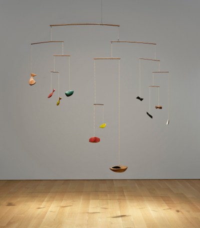 A mobile-like sculpture with abstract elements resembling boats and animals hangs from the wooden floor.
