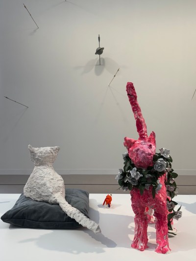 A sculpture of a pink cat wreathed with roses next to a sculpture of a white cat seated on a grey pillow.