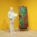 A man's white plaster sculpture, holding an easel, next to it is a painting of a feathered colored plant, leaning against two yellow walls.
