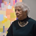 A woman stands next to an abstract painting of many superimposed colored squares.
