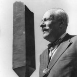 A man with a monocle hanging around his neck stands next to an abstract sculpture.