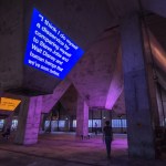 A person is projected against a blue screen with white text reading 