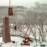 Bare trees in a snowy scene and a crane placing debris on an outdoor obelisk-shaped sculpture