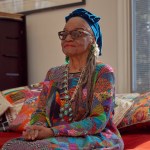 A black woman in a black turban and a colorful shirt sit on the sofa with patterned mats.