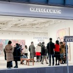 Scene of people in art gallery with paintings on the wall. There are signs in Chinese and English that say 