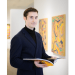 A clean-cut man wearing a dark shirt and blazer stands in an art gallery, holding a book.