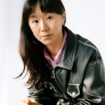 A long-haired woman with black bangs rests her elbows on her knees, clasping her hands, wearing a black leather jacket with pink buttons inside, and a serious expression