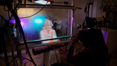 A white-haired woman smiles behind stained glass as someone takes a picture of her