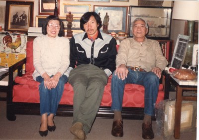 Three Asian Americans are sitting on the couch at home. An old woman sits on the left, an older man sits on the right, and between them is a young man in jeans. They live in a richly decorated house.