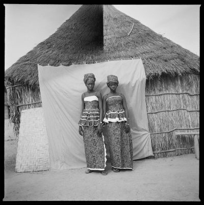 Two woman standing against a photo backdrop hung before a thatched structure.