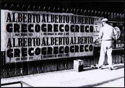 A man pasting a poster to a wall that features the words 'ALBERTO GRECO' over and over.