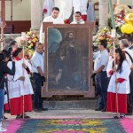 Two men hold up a large painting of St. Francis of Assisi in monastic robes during a ceremony at a church in Mexico, with church officials standing in the background and members of the public and two church workers in the foreground holding a menorah.