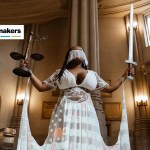 A black woman wears a white dress emblazoned with an American flag and has a white veil covering her face. She holds a scale and a sword.