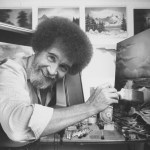 Black and white photo of a bushy-haired man looking at the camera, holding a paintbrush and leaning toward the canvas