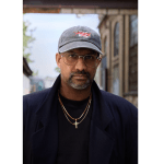 A young man wearing glasses, a baseball cap and a cross chain.