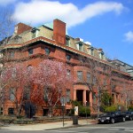 At the corner stood a stately building, dark red in color with a mansard roof. The nearby trees were bare except for a few with pink flowers. It's a sunny day.