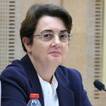A woman sits in front of a water bottle and a microphone.