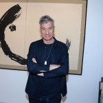 A white man stands with arms crossed in front of an abstract painting.