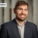 A man wearing a dark gray blazer and gray shirt smiles at the camera.