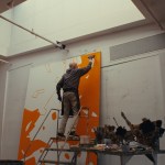 An image of a man standing on a ladder near the top of a large orange and white painting.