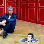 A photo of artist Maurizio Cattelan sits next to a statue of himself in a hole in the floor.