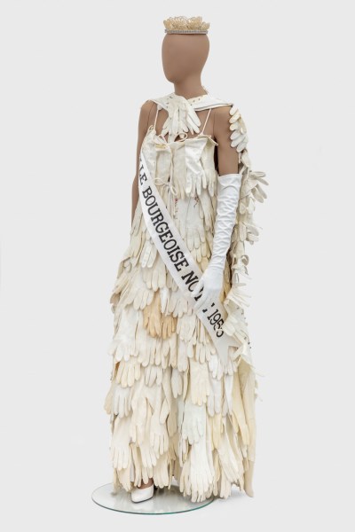 A brown mannequin wearing a dress made of many pairs of gloves. The dress's band reads