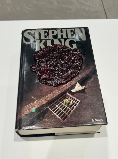 A Stephen King novel, with a storm drain on the cover, sits on a white pedestal in the gallery, with a dab of fruit spread piled on the cover.