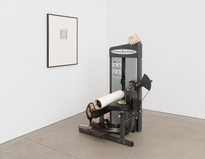 An artwork consists of many objects displayed together, most notably a weightlifting machine. Other items are placed on a weightlifting machine, and a framed piece of art hangs on the wall.