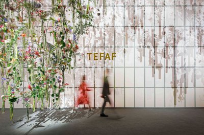 Two vague figures walked past a wall with TEFAF written on it. To the left is a hanging installation containing a variety of flowers and other plants.